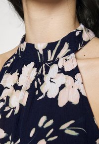 Niebieska bluzka na szyi z kwiatowym wzorem w odcieniach różu i zieleni. Materiał jest lekki, o subtelnej teksturze i fałdach przy dekolcie.