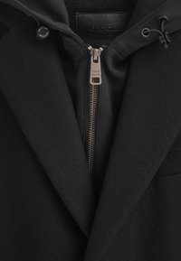 Svart kappa med en strukturerad yta, en dold huva och dragkedja med metalliska detaljer. Lappen säger "POLO CLUB."