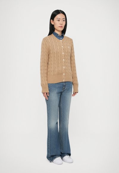 Bēša kabeli pītais kardigans ar apaļu kaklu un pogām, kombinēts ar zilām džinsa izplestām biksēm un baltām auduma kurpēm. Gluda tekstūra un piegulošs dizains.