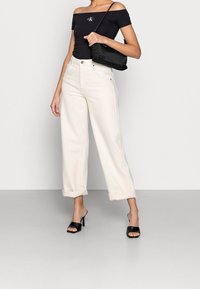 Svart off-shoulder top med CK-logotyp, vita höga vida jeans, svart handväska och svarta klackiga mule-skor.