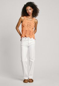 Oranje gehaakte tanktop met geschulpte zoom, gedragen met witte high-waisted jeans en bruine sandalen. Achtergrond is neutraal.