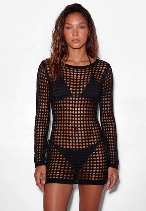 Donna che indossa un vestito nero a maniche lunghe a maglia aperta sopra un bikini nero, in piedi davanti a uno sfondo bianco uniforme.