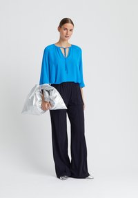 Blauwe blouse met een keyhole-halslijn, gecombineerd met vloeiende marineblauwe broek. Een zilveren tas en gestructureerde schoenen maken de outfit compleet. Glad stoffen texturen.