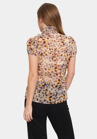 Transparente, kurzärmelige Bluse in Beige mit einem braun-blauen Punktemuster, Rüschensaum und leicht tailliertem Schnitt. Getragen mit schwarzen Hosen.