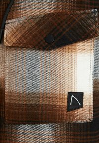 Tela de cuadros marrón y beige con un bolsillo que presenta un botón a presión negro. Incluye una etiqueta negra con un logo blanco. Textura suave.