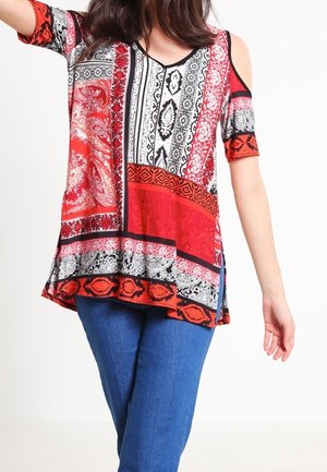 Woman wearing a red, black, and white patterned cold-shoulder top paired with blue jeans, standing against a plain background.