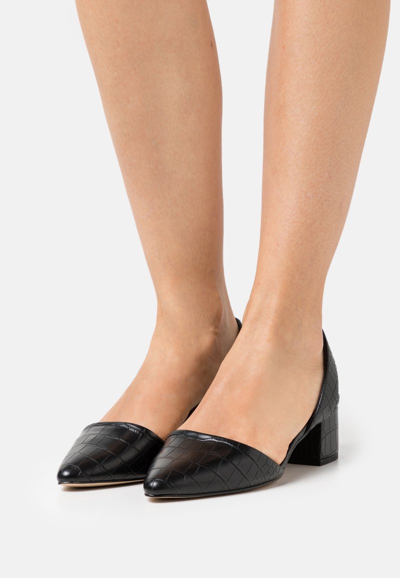 Zapatos de tacón de cuero sintético negro con punta afilada, patrón de cocodrilo texturizado y tacón cuadrado. El diseño presenta un escote bajo en el empeine.