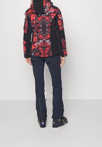 Kvinnors svart snöjacka med röd och svart abstrakt mönster; matchad med mörka byxor som har dragkedjor vid anklarna och svarta skidstövlar.