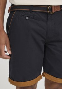 Marineblå shorts laget av lett materiale, med en brun rullet kant og kontraststing. Klassisk design med en knappelukket frontlomme.