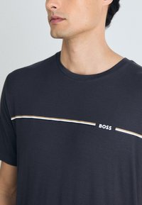 Tricou din bumbac de culoare gri închis, cu guler rotund, având un model de dungi orizontale în alb și bej, și un logo pe piept.