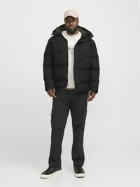Sort puffjakke med hætte, der har en grøn indvendig foring. Båret over en lys sweater og sorte cargo-bukser med lynlåse. Sorte sneakers.
