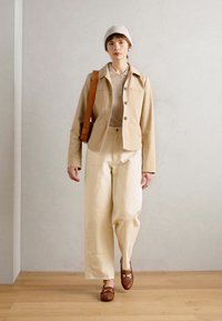 Bež jakna, kremasti majica i široke kremaste hlače. Smeđe cipele s naglascima. Model nosi svijetlu pletenu beanie kapu. Minimalistička postavka.