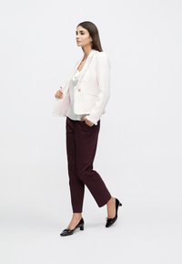 Veste en tweed rose clair avec bordure à franges, portée sur un haut crème et un pantalon ajusté bordeaux. Chaussures noires à petit talon avec un détail nœud.