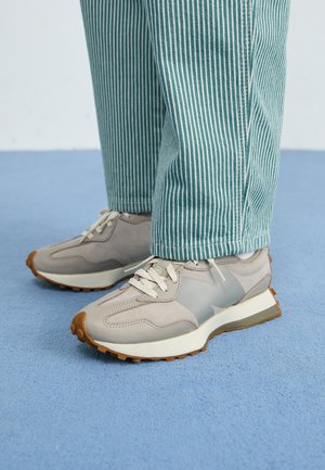 Baskets beige et grises avec des lacets blancs, portées avec un pantalon rayé verticalement bleu sarcelle et blanc, debout sur un sol moquetté bleu clair.