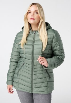 Vrouw met lang blond haar die een saliegroene doorgestikte capuchonjas en grijze broek draagt, staand tegen een lichte achtergrond.