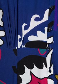 Gros plan sur un tissu bleu avec des motifs abstraits blancs, noirs, roses et turquoise, ainsi que de petits boutons métalliques en forme de cœur le long d'une couture.