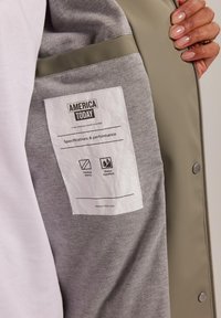 Harmaa tekstiili valkoisella lapulla, jossa lukee "AMERICA TODAY". Lapussa on tietoja lämpökohdistetuista saumoista ja vedenpitävistä ominaisuuksista, joka on sijoitettu vaalealle materiaalille.