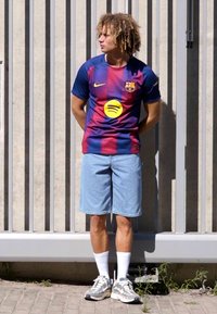Tricou FC Barcelona cu dungi albastre și roșii; pantaloni scurți din denim; șosete albe; pantofi sport cu model; stând în fața unei pereți știrb gri.