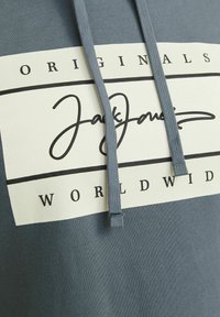 Grijze hoodie met een trekkoord, voorzien van een groot wit rechthoekig logo met zwarte tekst "Jack Jones Originals Worldwide." Gladde textuur.