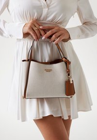Textured white handbag with brown accents, featuring a curved top, dual handles, and a decorative tag. Brand name "Guess" in gold.