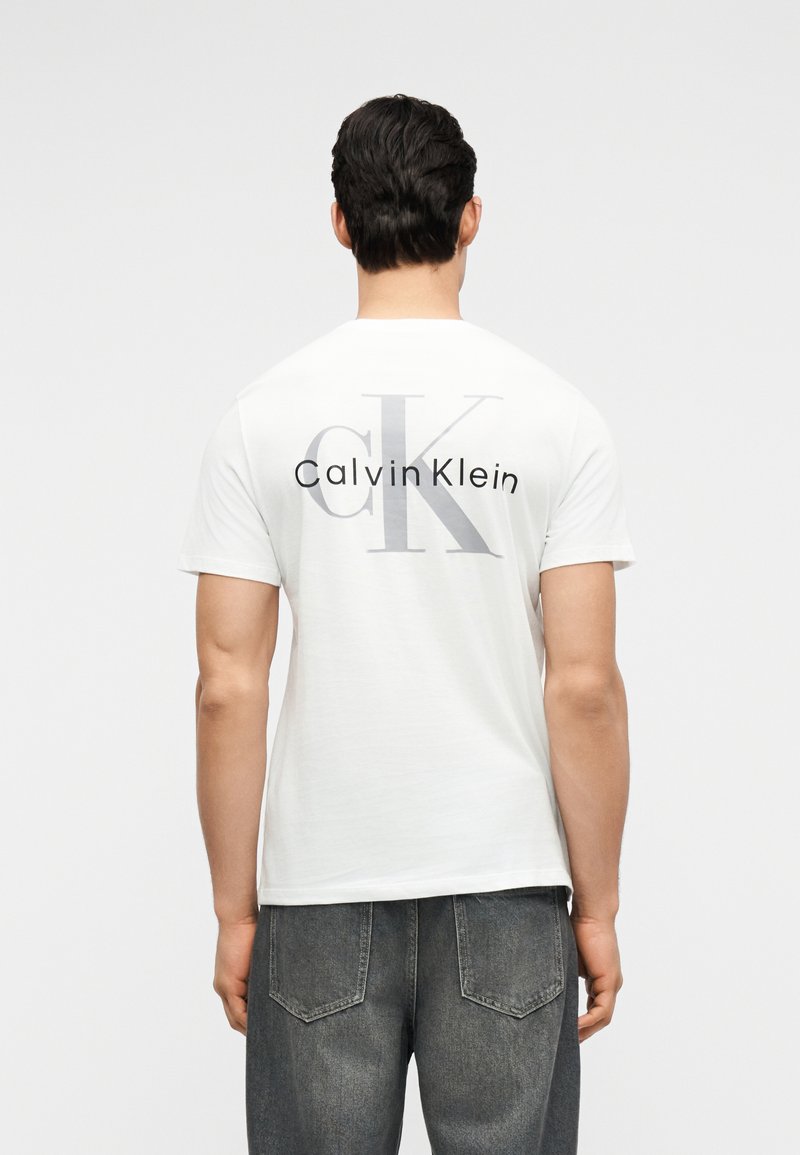 Άνδρας φαίνεται από πίσω, φοράει λευκό T-shirt Calvin Klein με μεγάλο γκρι και μαύρο λογότυπο "cK Calvin Klein" και γκρι τζιν.
