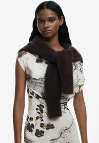 Sciarpa di lana marrone appoggiata su un vestito a fantasia con tonalità crema, nero e beige. La sciarpa presenta una texture morbida e soffice.