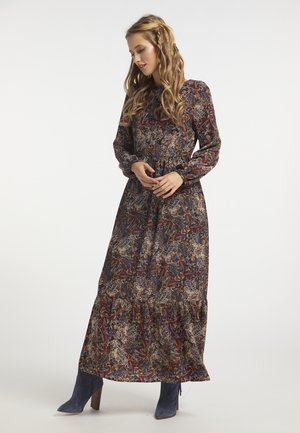 Vrouw met lang golvend haar, gekleed in een lange paisley maxi-jurk met lange mouwen en blauwe enkellaarsjes, staand met de handen in de taille gevouwen.