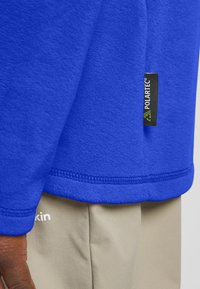 Blauwe fleece trui met een Polartec-label, met een zachte textuur en gestikte zoom, gedragen over lichtbeige broeken met een logo.