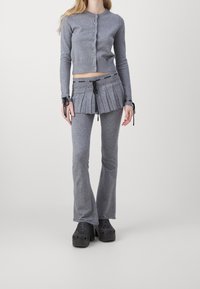 Szary sweter robiony na drutach z guzikami, zestawiony z plisowaną szaro-skalną spódnicą z troczkami i ozdóbkami, oraz szarymi spodniami w dzwony. Czarne klompy.