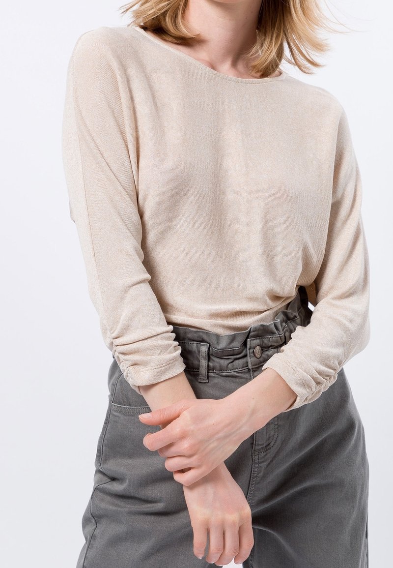 Personne portant un haut beige à manches longues et un pantalon gris taille haute avec taille froncée, mains croisées devant, recadré au-dessus du cou.