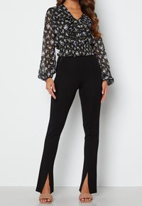 Blusa nera floreale con scollo arricciato, maniche lunghe trasparenti e vita elastica; abbinata a pantaloni neri attillati con orli a spacco.