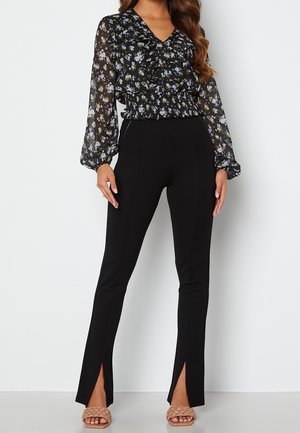 Blusa nera floreale con scollo arricciato, maniche lunghe trasparenti e vita elastica; abbinata a pantaloni neri attillati con orli a spacco.