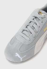 Baskets en daim gris clair avec des lacets plats, une marque dorée et un logo blanc. Présente un design texturé et un panneau latéral contrastant.