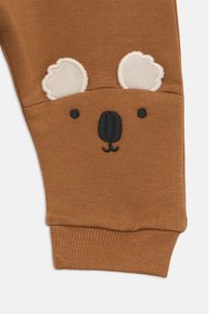 Καφέ πλεκτό ύφασμα με ραμμένο πρόσωπο κοάλα, που περιλαμβάνει μαύρη μύτη, μάτια και λευκά αυτιά, στην μανσέτα ενός ρούχου.