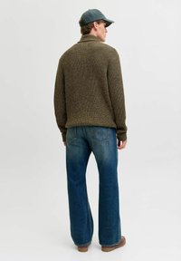 Uomo che indossa un maglione a maglia verde, jeans blu, scarpe marroni e un berretto grigio, in piedi con la schiena rivolta verso uno sfondo bianco semplice.