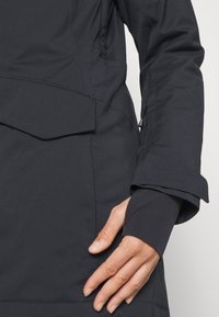 Veste imperméable noire avec une texture lisse, dotée d'un rabat de poche et de poignets avec trou pour le pouce. Conçue pour l'isolation et la protection contre les intempéries.