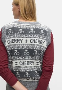 Szary dzianinowy zamek bez rękawów z białym wzorem rowerowym i napisem "CHERRY". Ściągacze na krawędziach, noszony na burgundowej bluzie z długim rękawem.