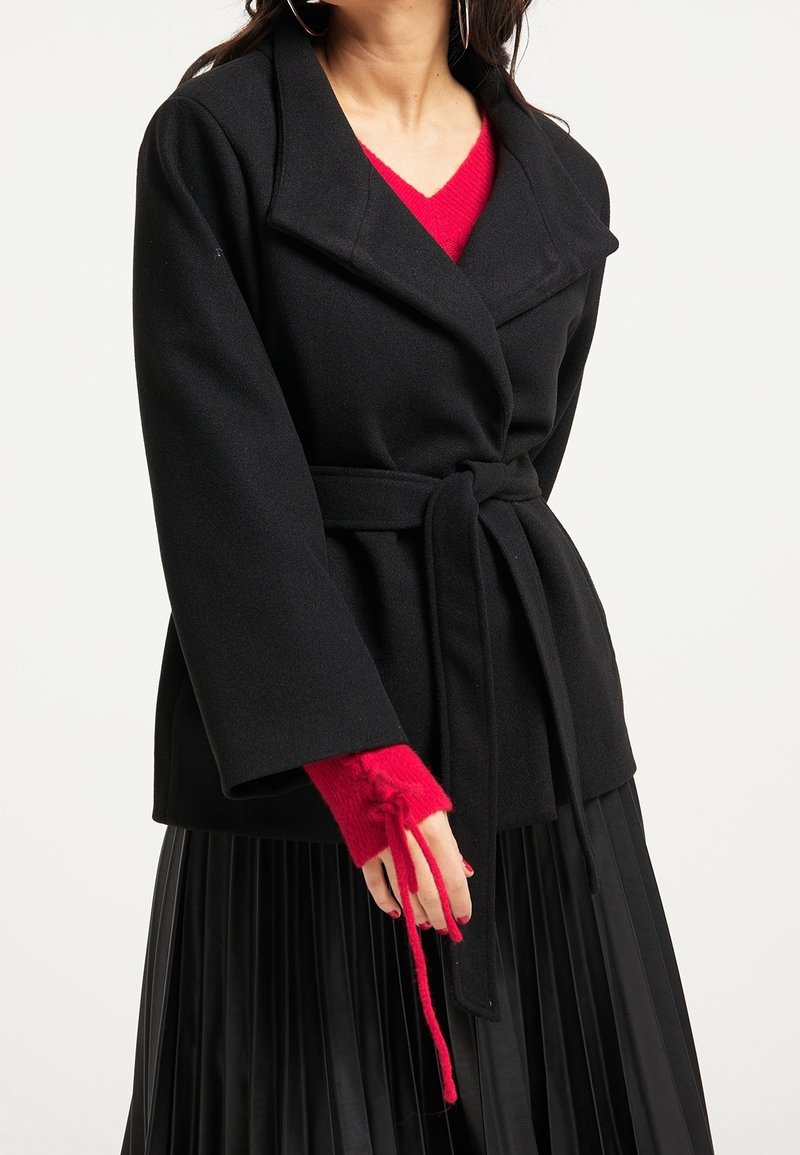 Manteau noir portefeuille avec col, taille ceinturée et manches larges, associé à un pull rouge avec des mitaines. Tissu texturé et design en couches.