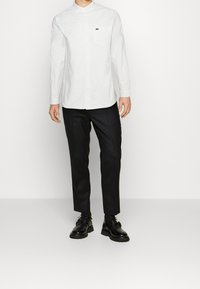 Man med vit långärmad skjorta med knapp stolpe och en liten logotyp på bröstet, svarta byxor och svarta skor, som står mot en vit bakgrund.