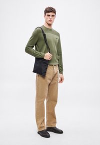 Fekete keresztpántos táska cipzáras záródással és állítható pánttal, zöld bordázott pulóverrel és barna, bő szárú nadrággal párosítva. Fekete cipők.