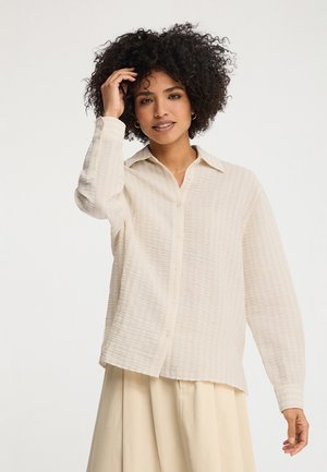 Vrouw met krullend haar, gekleed in een textuurcrèmekleurig overhemd met knopen en een beige rok, die haar haar aanraakt tegen een neutrale achtergrond.