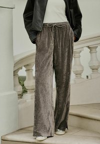 Grijze fluwelen wijde broek met een verstelbare tailleband en zijsplitdetails, gecombineerd met een getailleerde top en een zwarte jas.