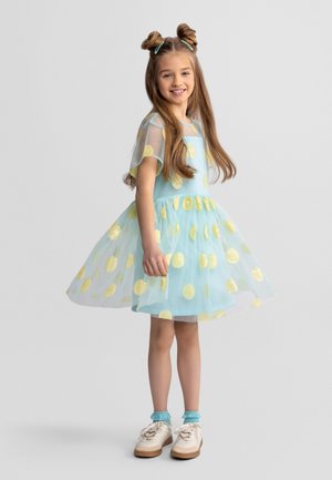 Junges Mädchen mit langem Haar im blauen Kleid mit gelben Punkten, blauen Socken und weißen Turnschuhen, lächelnd und leicht zur Seite gedreht.