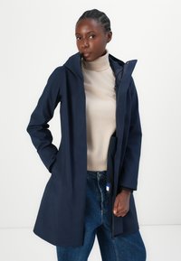 Manteau imperméable bleu marine avec capuche, doté d'une fermeture éclair et de poches latérales, porté sur un pull-over beige à col roulé et un jean bleu.