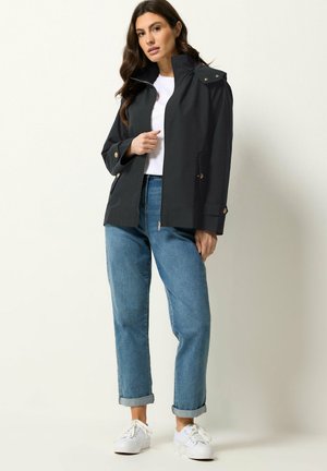 Kvinde iført en sort lynlåstrøje med knappedetaljer, hvid t-shirt, opsmøgede blå jeans og hvide sneakers, stående foran en ensfarvet baggrund.