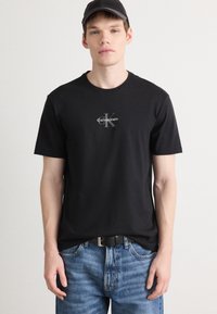 T-shirt de algodão preto com um logótipo cinza da Calvin Klein centrado no peito. O modelo combina-o com jeans de denim texturizado azuis.