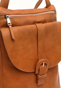 Smeđi kožni ruksak s preklopnom zatvaranjem, podesivim remenom i srebrnim zatvaračem. Glatka tekstura s ušivenim detaljima i naglaskom na kopču.