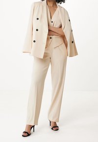 Béžový oblek na míru s dvouřadým sakem a černými knoflíky, doplněný o odpovídající kalhoty. Oblečené s černými vysokými sandály.