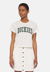 Weißes, verkürztes T-Shirt mit grüßen "DICKIES"-Aufdruck, runder Halsauschnitt und kurzen Ärmeln. Kombiniert mit einem weißen, knopfgeknöpften Rock.