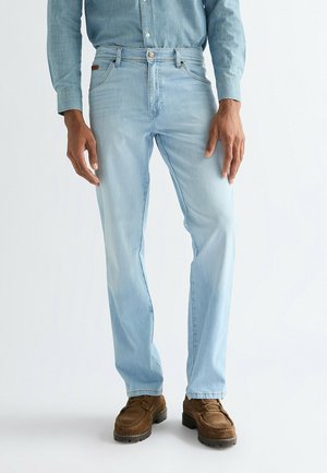 Mand iført lyseblå jeans, en indstøbt denimskjorte og brune ruskindsløve mod en ensartet hvid baggrund.